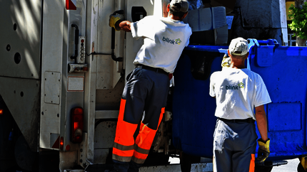 Twee vuilnismannen zijn bezig een blauwe container in een vuilniswagen te ledigen.