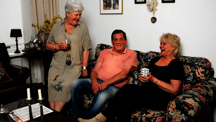 Foto van een woonkamer met een senioren stel op de bank en een vijftiger. Lachend.