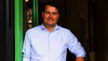 Foto van Dries Ewalds vanaf middel omhoog. Met lichtblauwe blouse en glimlach. In een deuroperning, leunend tegen groene deurstijl. 