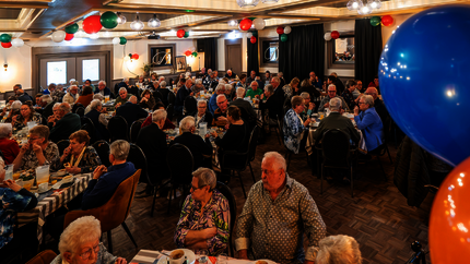 Overzichtsfoto van jubileumlunch met tientallen oudere huurders aan tafeltjes in een grote zaal. 