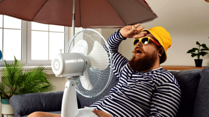 Man in een blauw wit gestreept shirt en geel hoedje zwetend op de bank met een rode parasol en ventilator