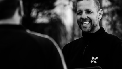 zwart wit foto van een medewerker van elk van de borst omhoog. Lachend. Met een zwarte trui met een wit elk logo.