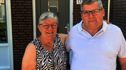 Jan en Francie in de zon voor hun gerenoveerde woning. Vanaf hun middel tot hoofd op de foto. Jan met witte polo. Francie met een zwart/witte mouwlos hes. Beiden dragen een bril.