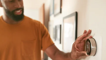 Een man met een donker huidskleur een baard en snor en oranje shirt draait aan de thermostaat in een woning. Met tekst 'Noodfonds Energie' op de foto.
