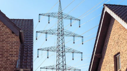 Twee daken en bovenste verdiepingen van huizen met daartussen blauwe lucht en een hoogspannignsmast met kabels.