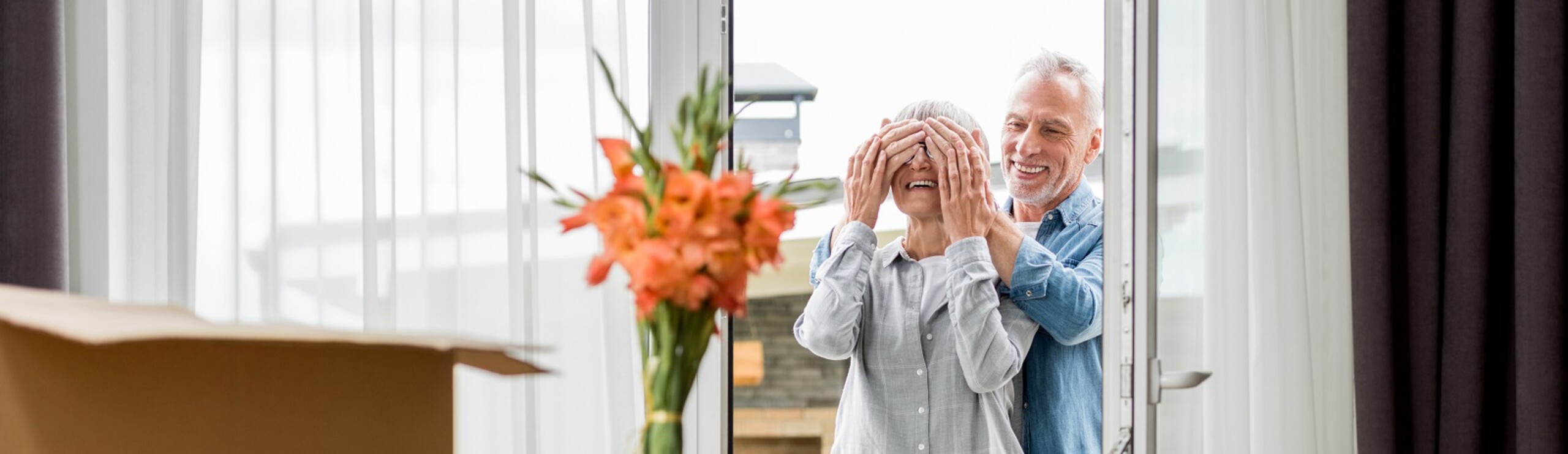 Ouder stel loopt door de voordeur. Man houdt zijn handen voor de ogen van de vrouw.