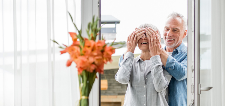 Ouder stel loopt door de voordeur. Man houdt zijn handen voor de ogen van de vrouw.